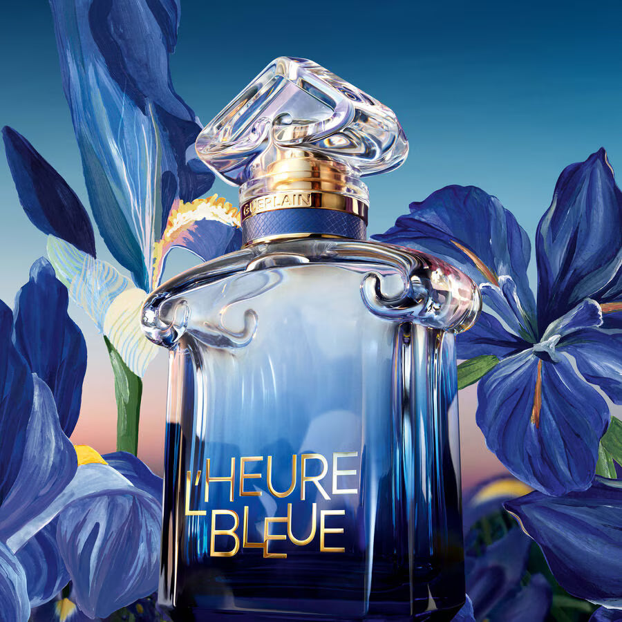 Guerlain The Hourglass perfume bottle in a rich blue glass, surrounded by large blue irises on a blue gradient background. The bottle features a clear faceted cap, a gold collar, and 'THE HOURGASS' and 'GUERLAIN' branding.