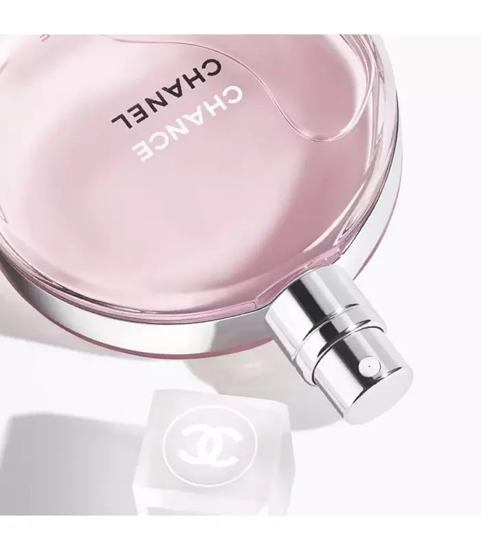 A close-up of a Chanel Chance Eau Tendre perfume bottle with a pink hue and a silver cap, alongside a white square object which appears to be a lid or packaging component.